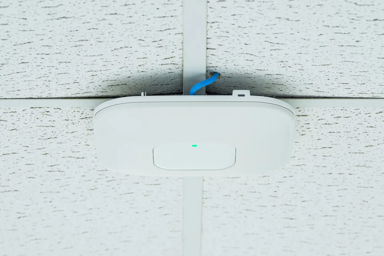 White wireless access point mounted on a drop ceiling grid, with a blue Ethernet cable feeding in from above and a green status light lit