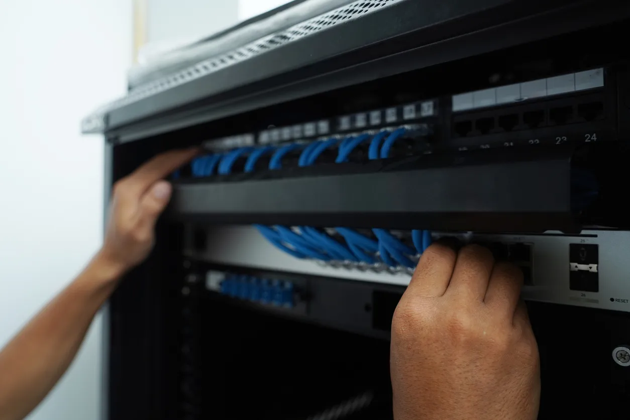 Two hands adjusting a network switch and patch panel in a server rack, with blue Ethernet cables run to numbered ports