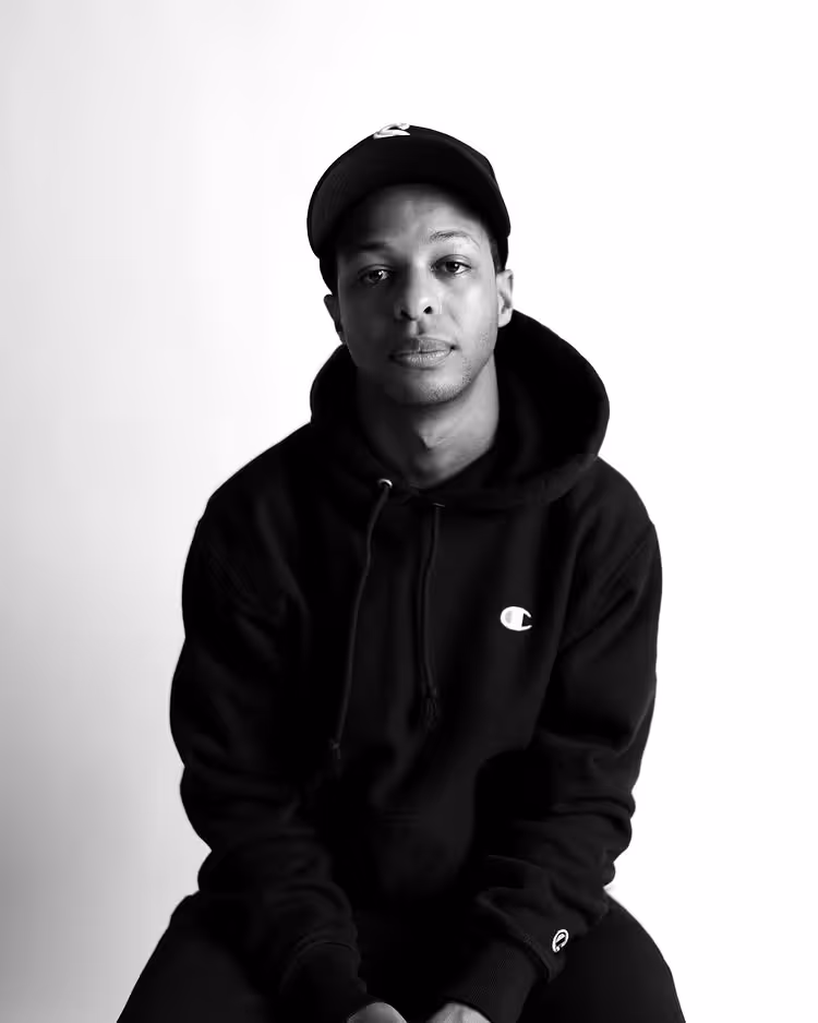 Man wearing a black hoodie and baseball cap sitting against a plain white background.