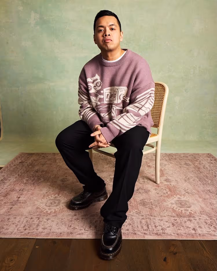 Man sitting on a chair wearing a purple sweater with graphic design and black pants on a rug.