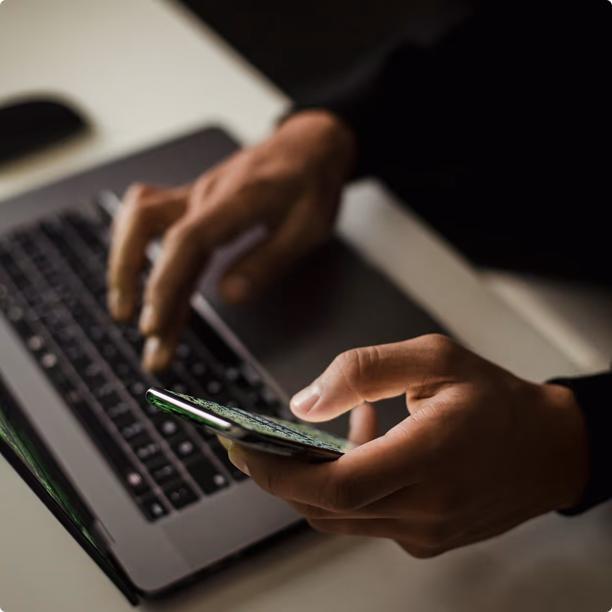 Person using smartphone with one hand and typing on laptop keyboard with other hand