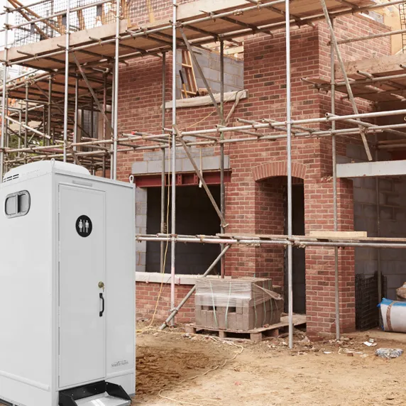 White portable restroom with ventilation window outside brick building under construction with scaffolding