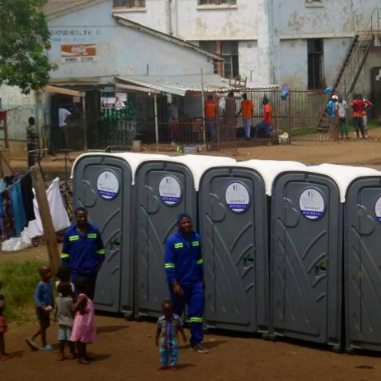 Row of gray portable restrooms with white roofs outdoors near building and people walking around