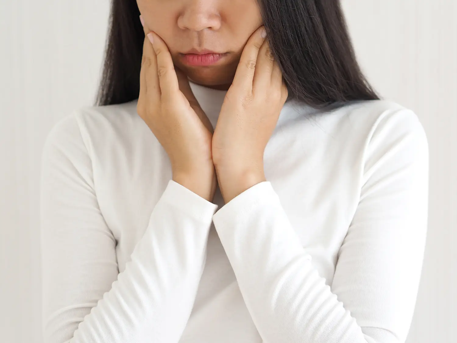 Person in white long sleeve holds both cheeks with hands, showing discomfort or pain.