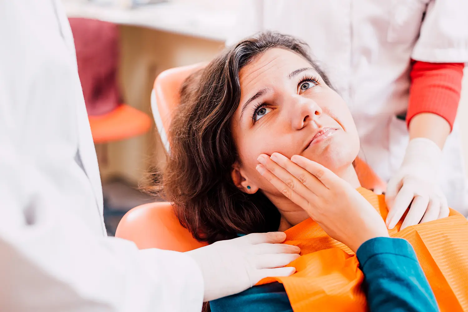 Woman in dental chair holding jaw in pain while two gloved dental professionals assist her.