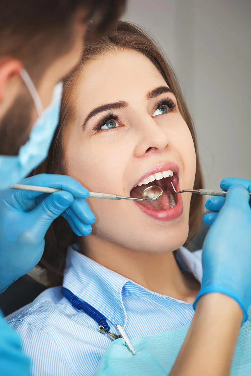 Dentist wearing gloves and mask examining a woman's open mouth with dental tools.