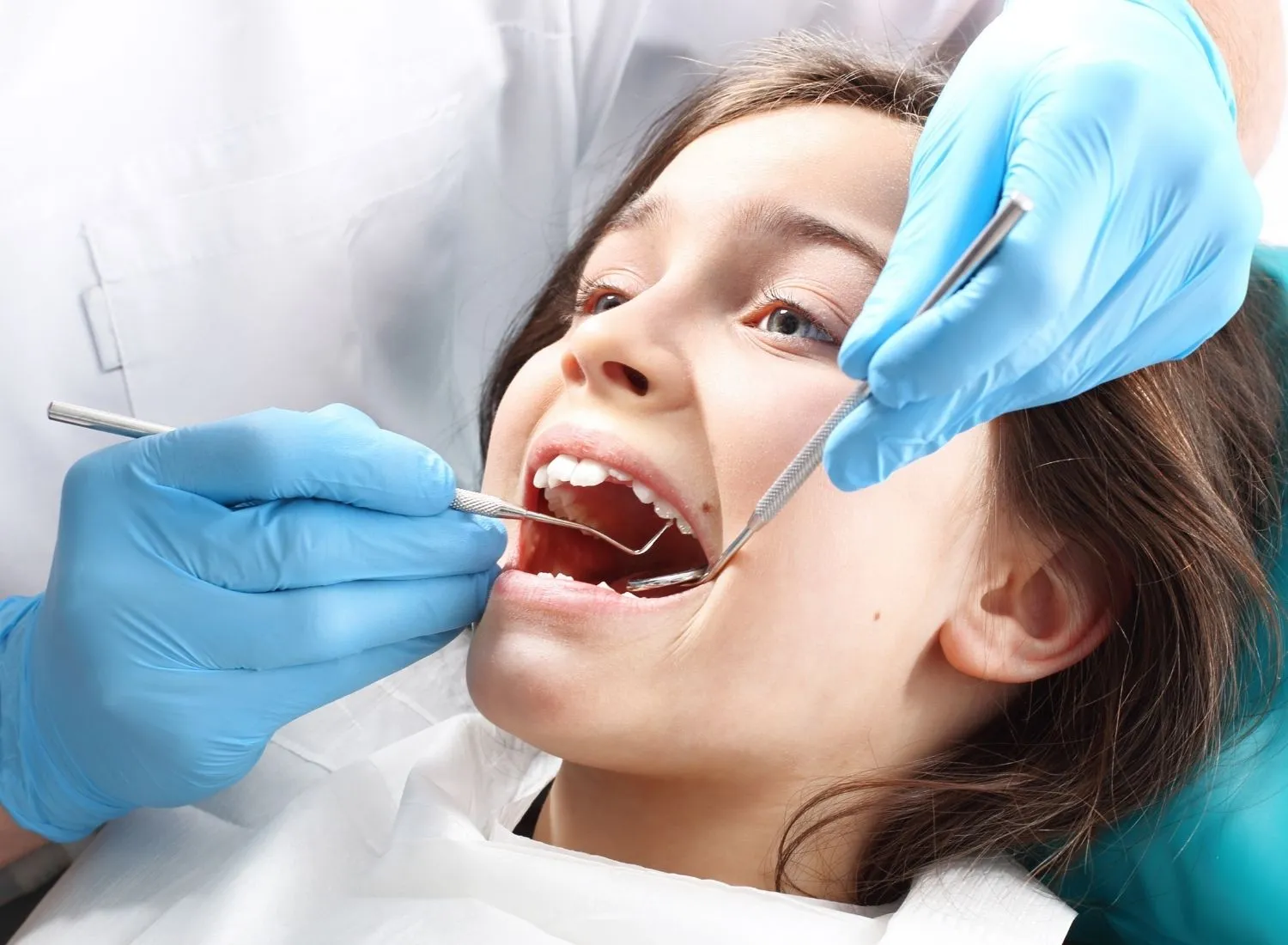 Young girl with mouth open during dental checkup by a dentist wearing blue gloves and holding dental tools.