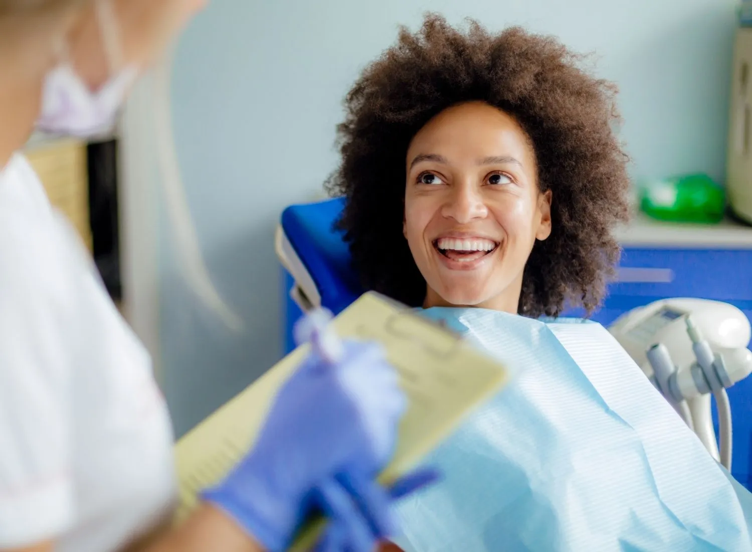 Smiling woman in dental chair talking to dentist holding a clipboard and wearing blue gloves.