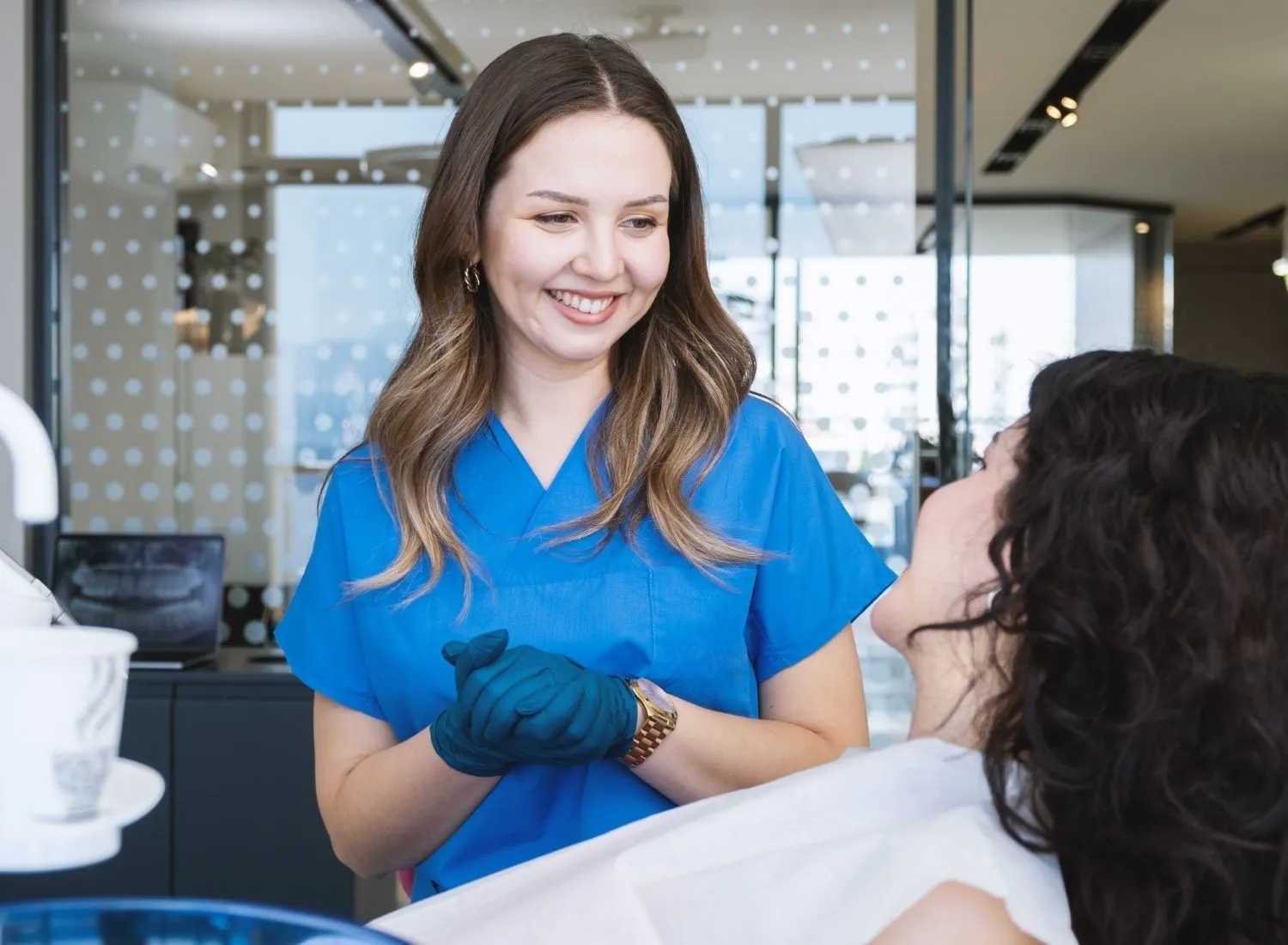 Smiling female dental professional in blue scrubs talking to a reclining patient in a dental office.