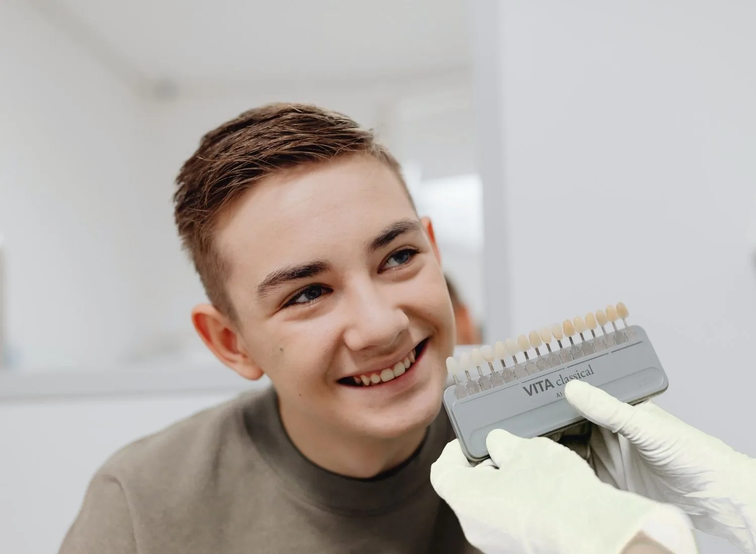 Smiling young man looking at dental shade guide held by gloved hands in a dental office.