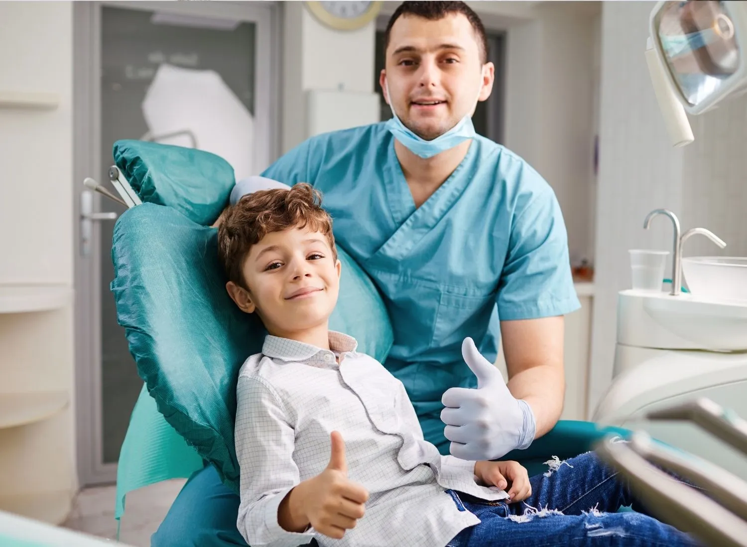 Boy sitting in dentist chair and dentist in blue scrubs giving thumbs-up in clinic.