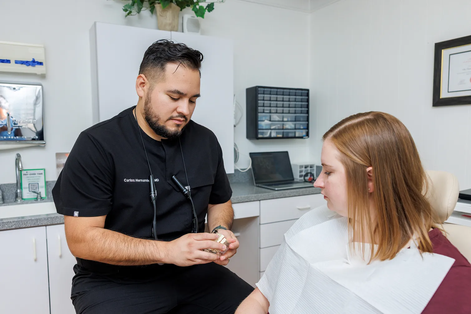 Male healthcare professional in black scrubs with name tag 'Carlos Hernandez MD' seated next to a seated female patient wearing a dental bib, holding a dental model while explaining.