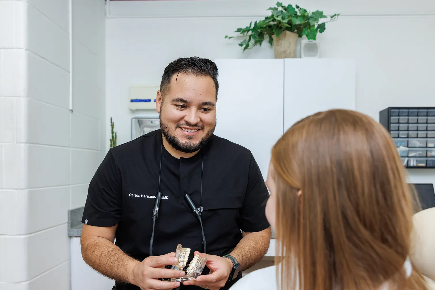 Smiling male dentist in black scrubs holding a dental model while talking to a female patient with red hair in a dental office.