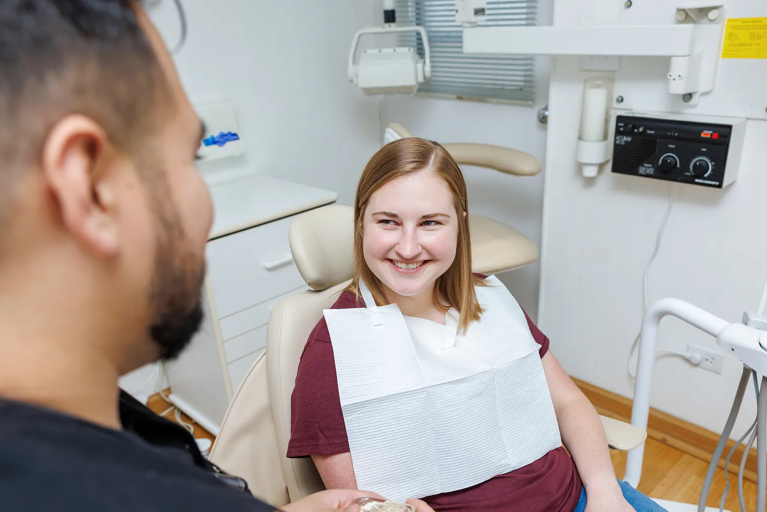 Smiling woman sitting in a dental chair wearing a dental bib, looking at a dentist in a dental office.