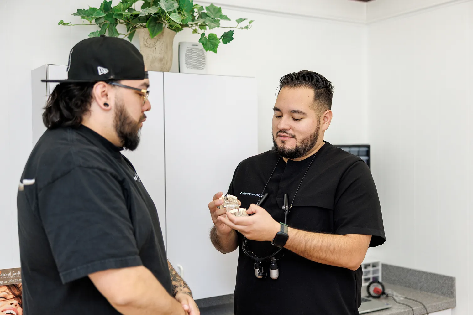 Two men in a dental office; one man wearing a backward cap and glasses listens while the other, wearing dental loupes and a name tag, holds and shows dental models.