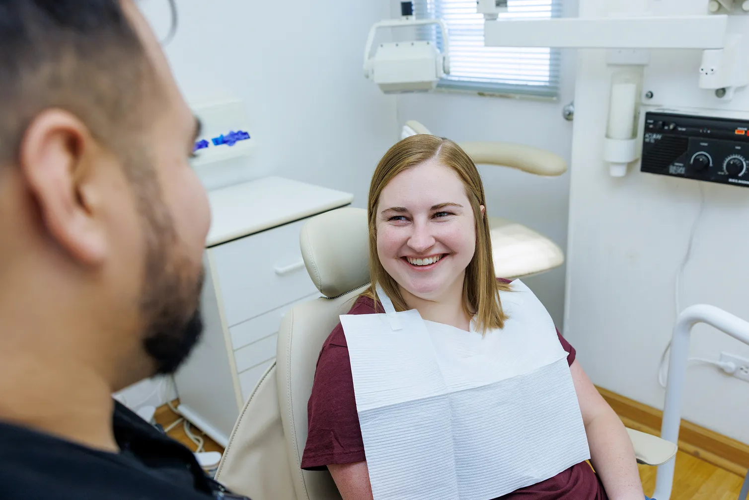 Smiling woman sitting in a dental chair wearing a dental bib, looking at a person partially visible in the foreground in a dental clinic.