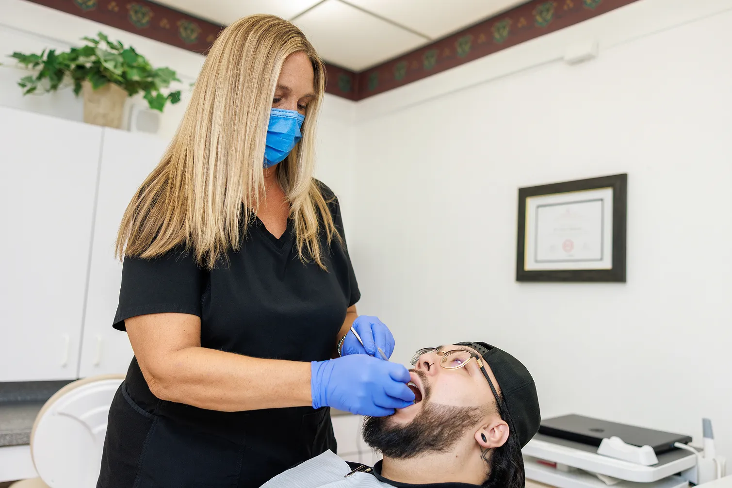 A female dental professional wearing a blue mask and gloves examining the open mouth of a male patient with glasses and a black cap in a dental office.