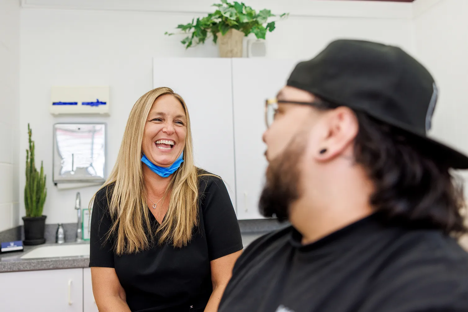 A smiling woman with long blonde hair and a lowered blue face mask talks with a man wearing a black cap and glasses in a white room with cabinets, a plant, and a sink.