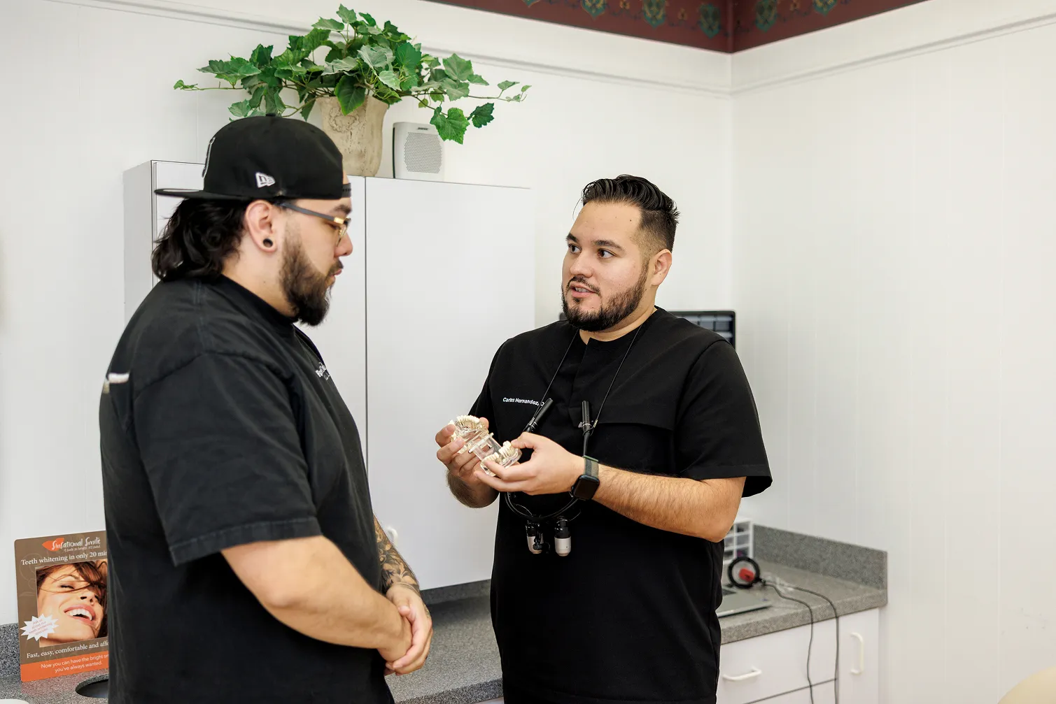 Two men in a clinical setting; the man on the right wearing black scrubs and holding a dental model is explaining something to the man on the left wearing a black t-shirt and backwards black cap.