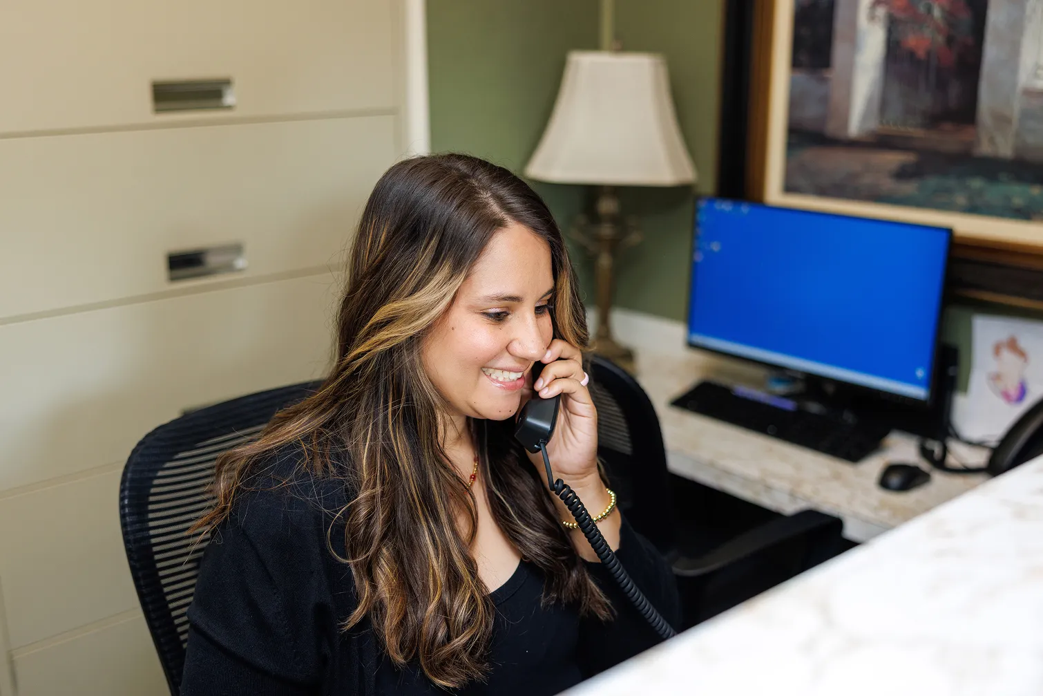 Smiling woman with long highlighted hair sitting at an office desk, talking on a corded phone, with a computer monitor and lamp in the background.