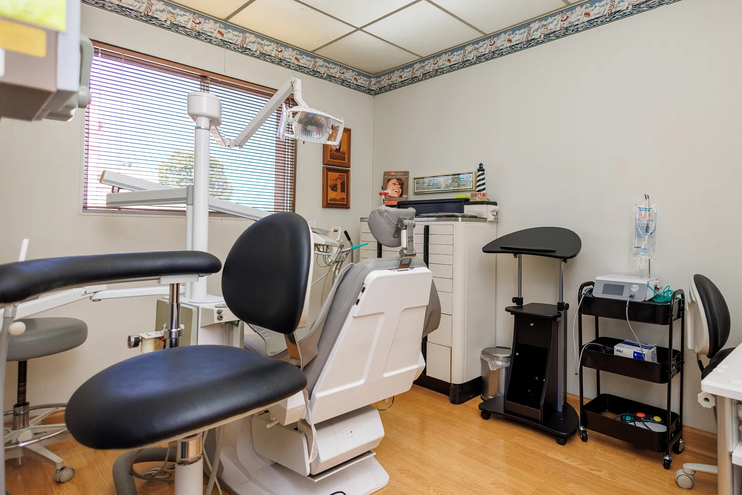 Interior of a dental office showing a dental chair with attached dental light and instrument tray, black stools, a window with blinds, cabinets with dental supplies, a black mobile cart, and a white mobile cart with medical equipment and IV fluid bag.