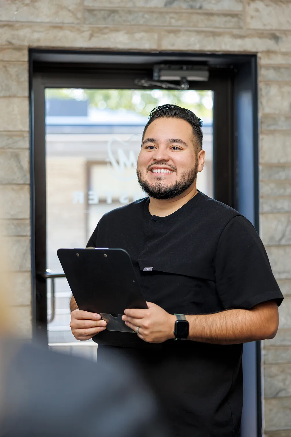 Smiling man with a beard and mustache, wearing black scrubs, holding a clipboard, standing in front of a glass door with a stone wall background.
