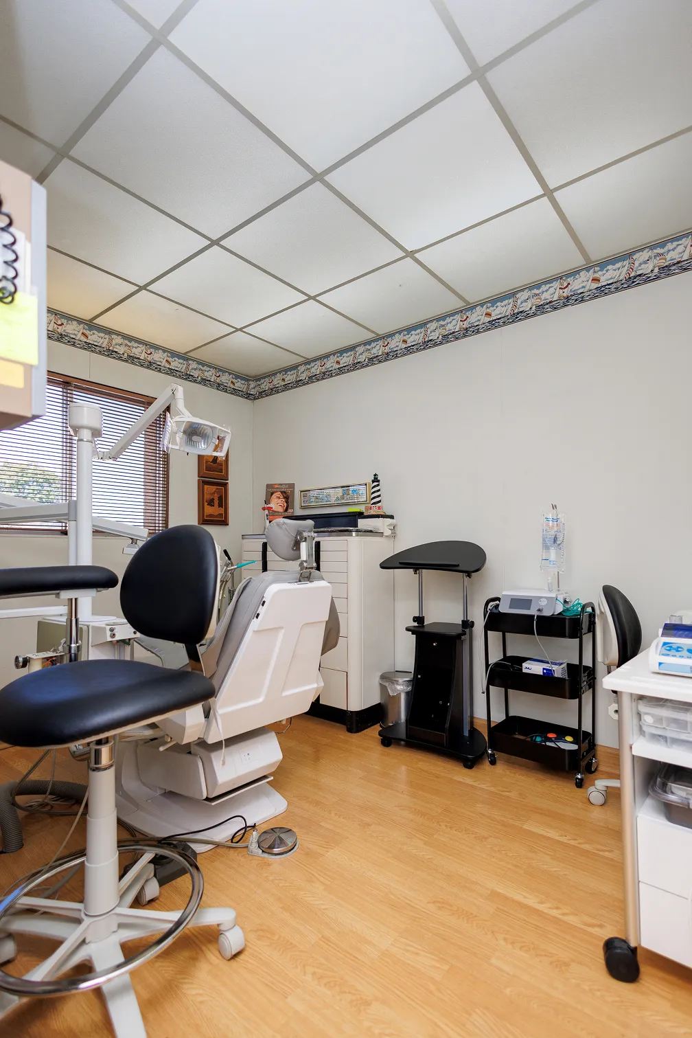 Interior of a dental clinic room with a dental chair, a dentist stool with black seat, a dental light, cabinets, and medical equipment against white walls and wood-look flooring.
