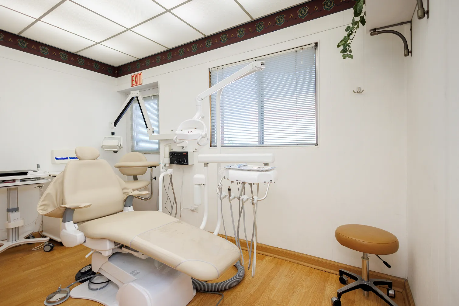Dental examination room with beige dental chair, dental instruments mounted on a movable arm, beige stool on wheels, and windows with blinds, featuring wood flooring and white walls.