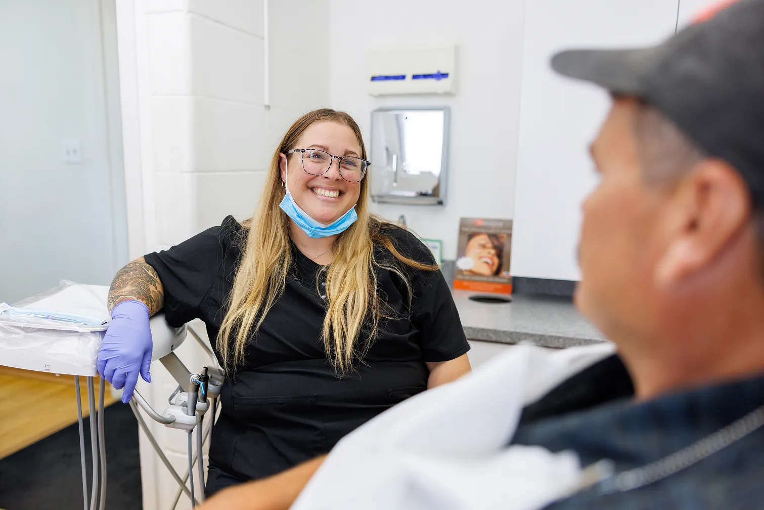 Smiling female dental professional with long blonde hair, glasses, and a blue disposable mask under her chin, sitting and leaning on dental equipment, talking to a patient in a dental office.