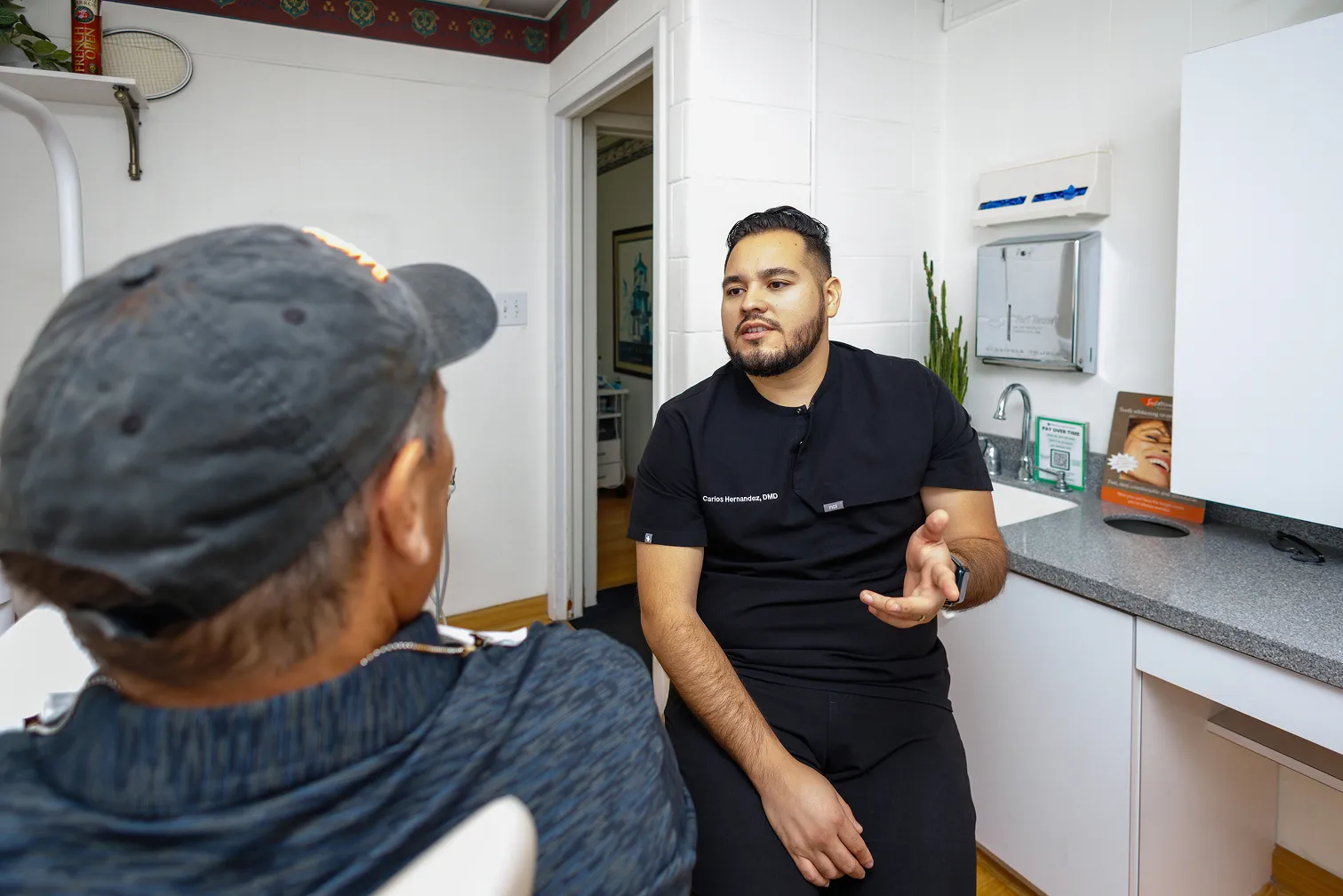 A dentist wearing black scrubs with the name Carlos Hernandez, DMD, talks to a patient seated in a dental chair, inside a dental office with white walls and cabinets.