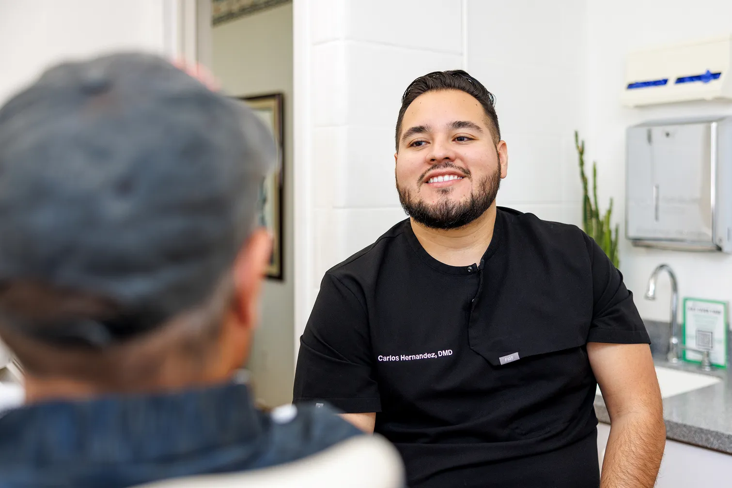 Smiling male dentist wearing black scrubs with name tag 'Carlos Hernandez, DMD' talking to an elderly patient in a clinical setting.