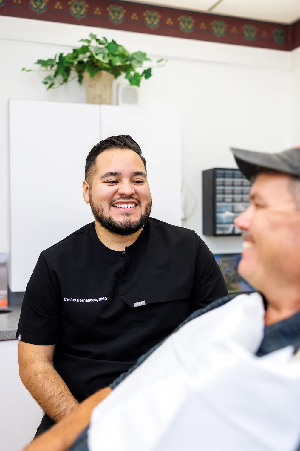Smiling male dentist in black scrubs with name tag 'Carlos Hernandez, DMD' talking to a seated male patient wearing a dental bib, in a dental office with cabinets and a potted plant in the background.