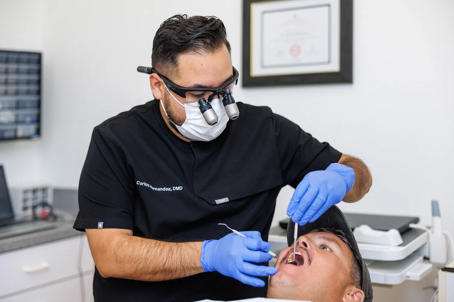 Dentist wearing a black scrub top, face mask, and magnifying loupes examining a patient's open mouth using dental tools, in a clinical setting.