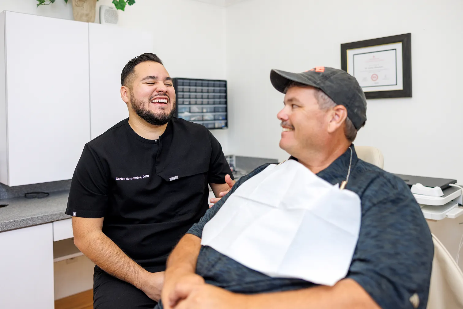 Smiling dentist in black scrubs with name tag 'Carlos Hernandez, DMD' interacts happily with a male patient wearing a dental bib and cap seated in a dental chair in a clinical room.