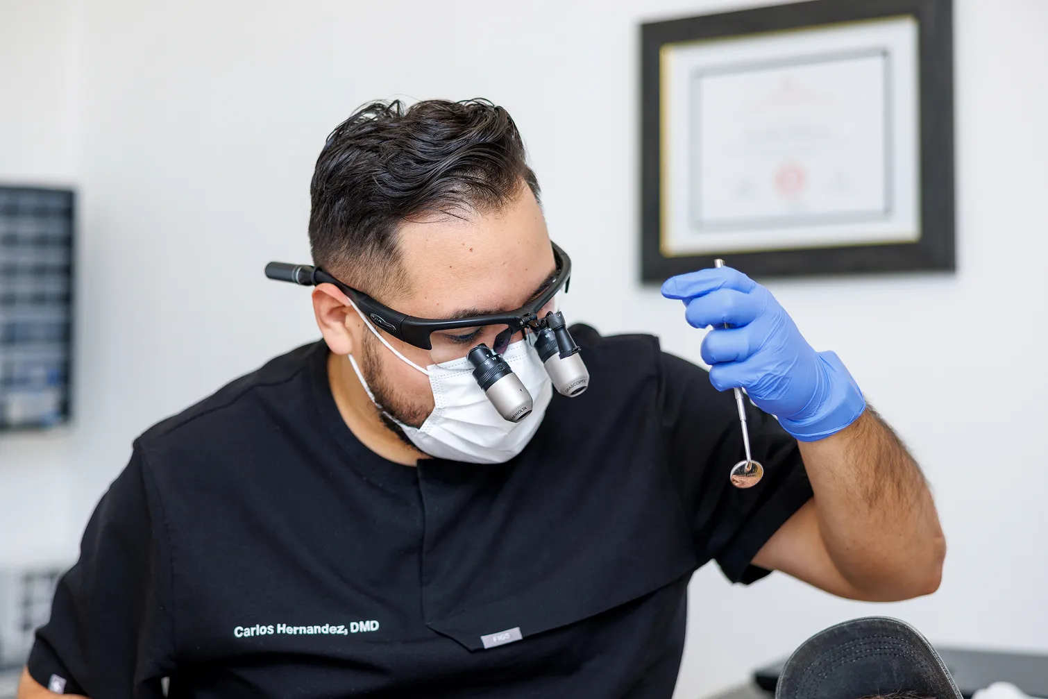 Dentist wearing magnifying glasses, a mask, and blue gloves examining a patient using a dental mirror.