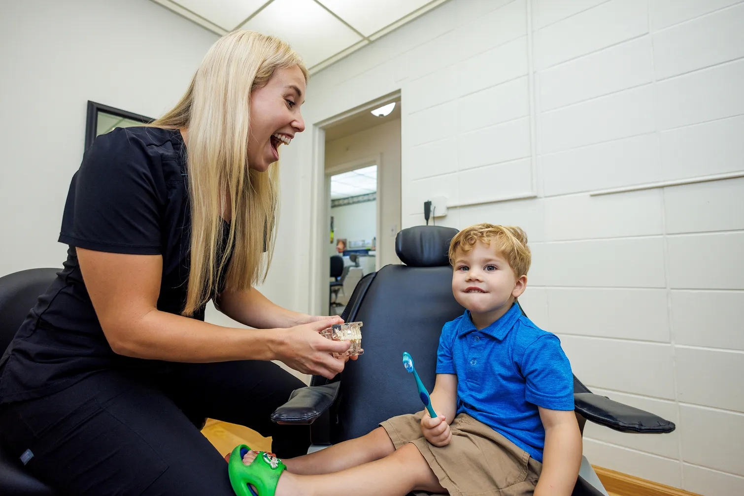 A female dental professional in black scrubs smiles and talks to a young boy sitting in a dental chair holding a blue toothbrush, in a bright dental office.