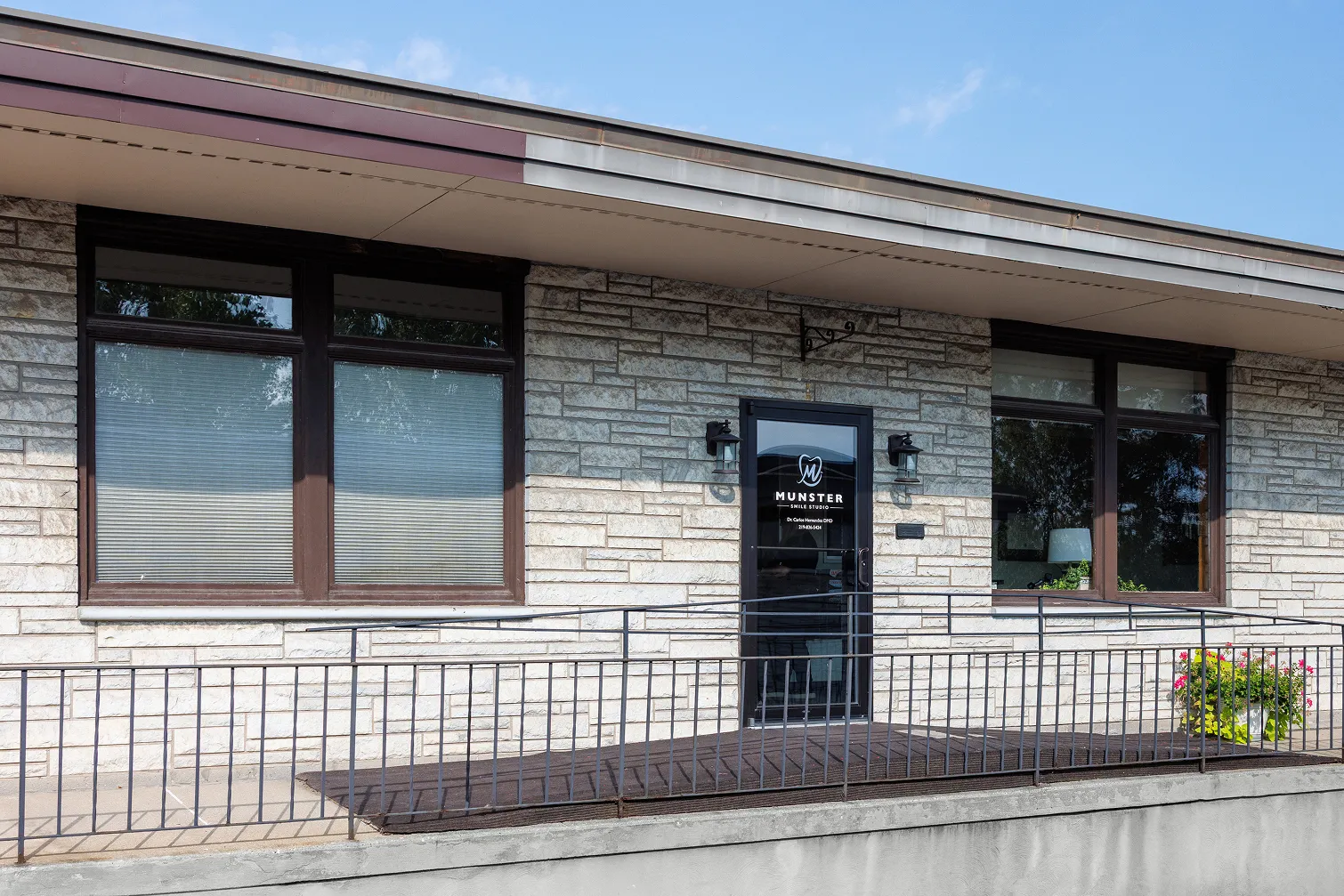 Entrance to Munster Smile Studio with a black door displaying the logo and name, flanked by two black wall lamps, set in a stone exterior wall with two large windows and a metal railing in front.