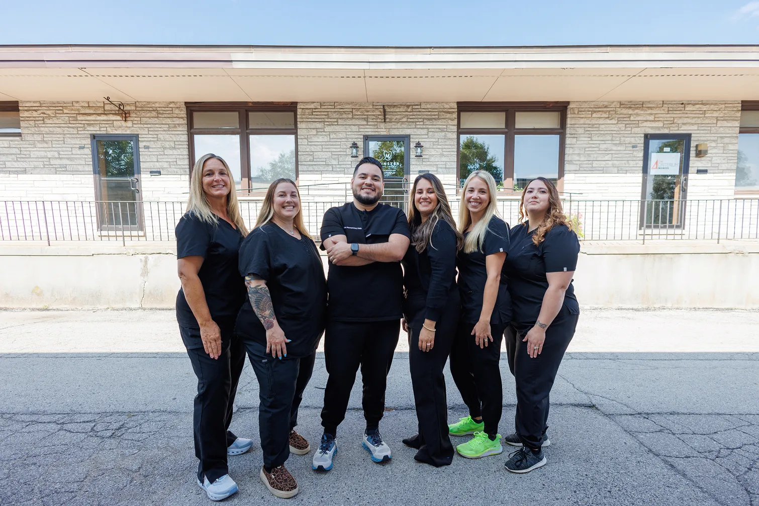 Group of six smiling healthcare professionals standing outside in front of a building with large windows and stone walls, all wearing black scrubs with varied casual shoes.