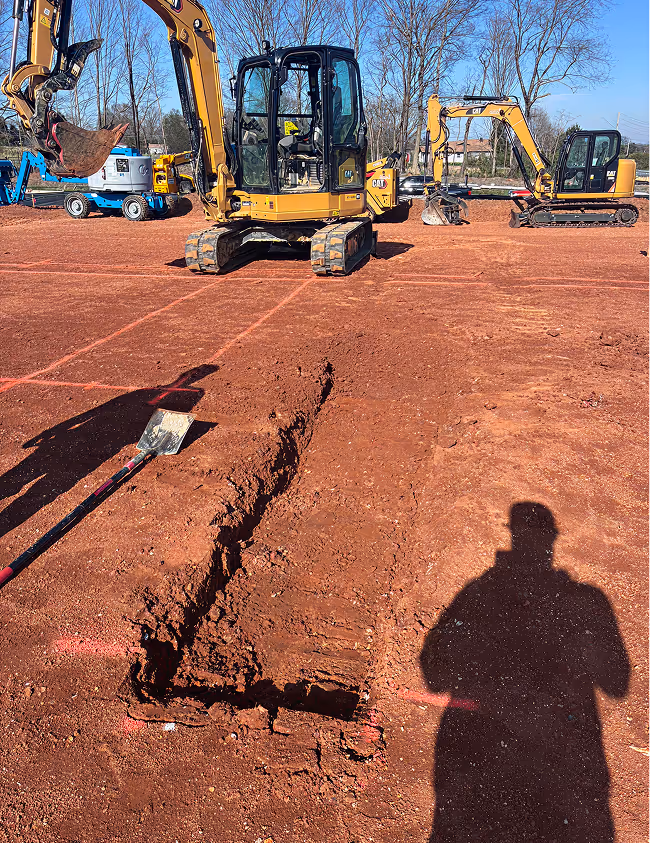 Construction site with excavators, a trench dug in red soil, and a shovel on the ground.