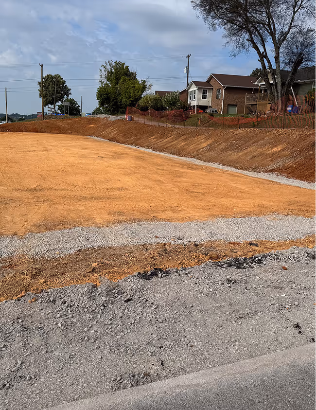 Cleared construction site with dirt and gravel near residential houses under a cloudy sky.