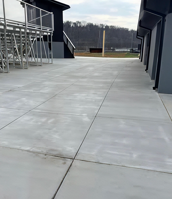 Concrete pavement between two buildings with metal bleachers on the left and trees in the background.