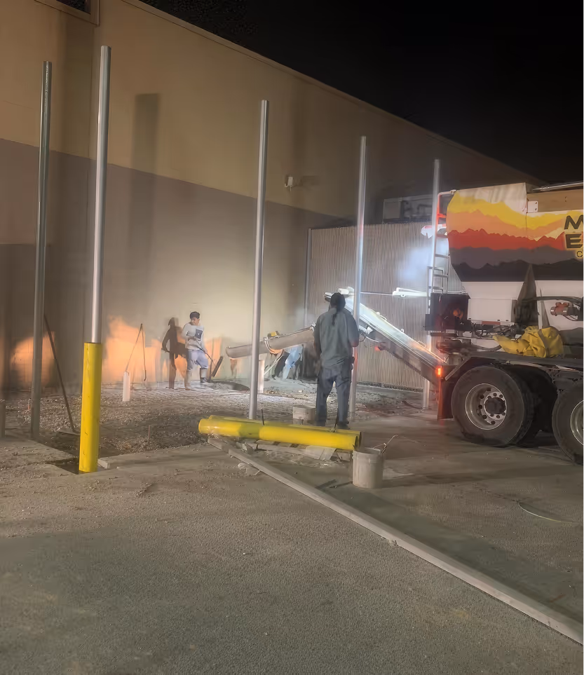 Workers pouring concrete at night near metal poles and a truck with a colorful design.