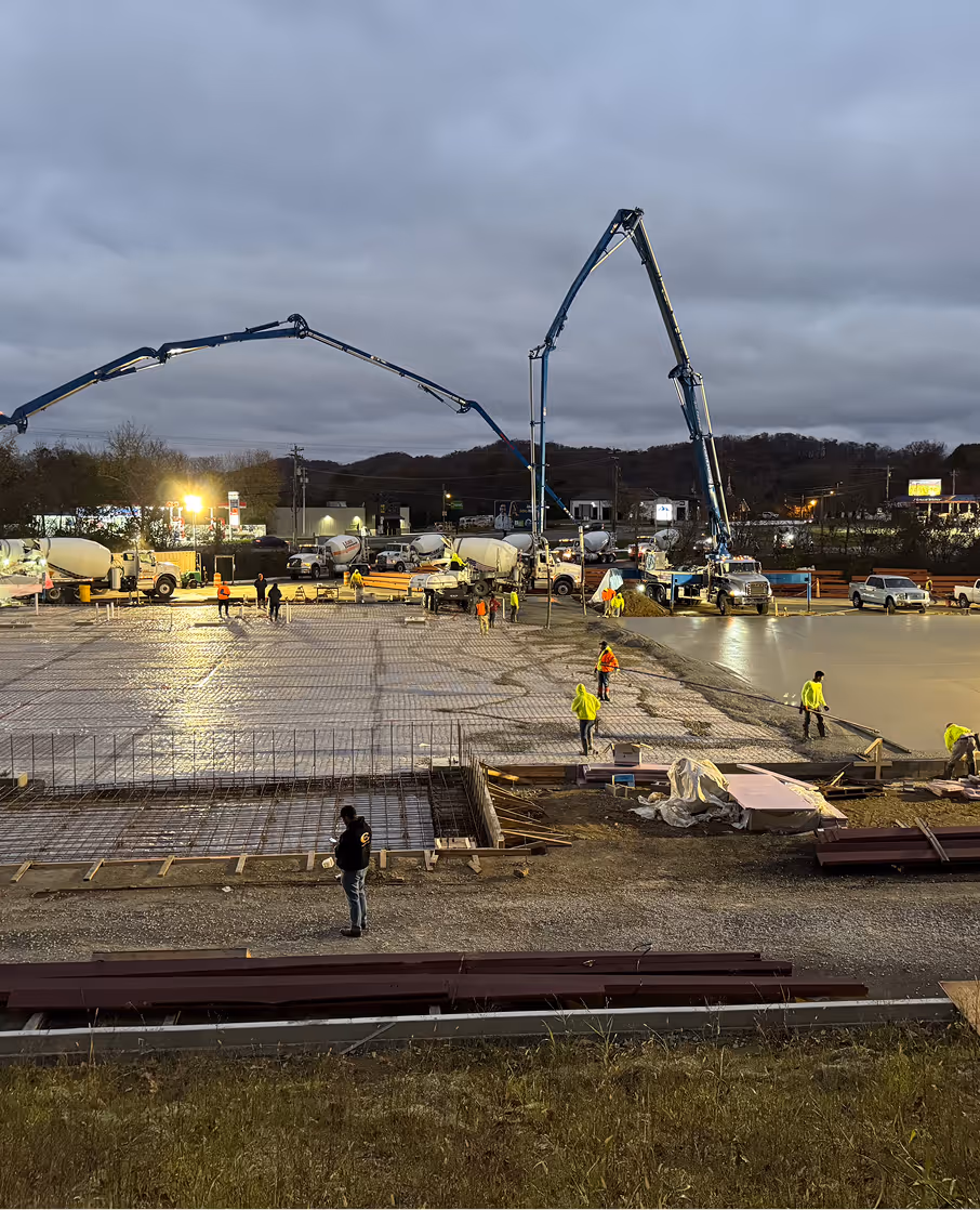 Construction site at dusk with workers pouring and smoothing concrete using machinery and tools.