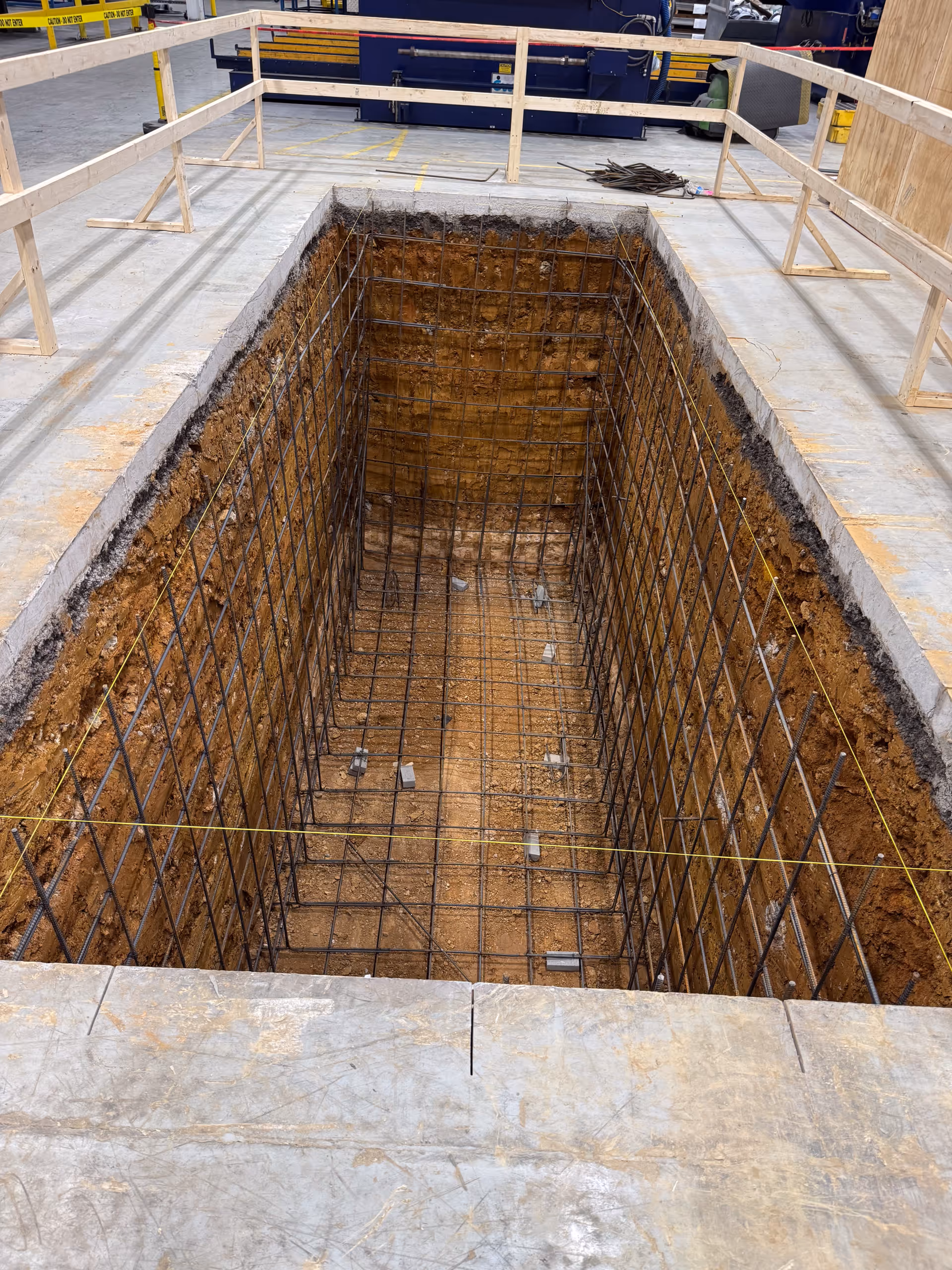 Deep rectangular construction pit with steel rebar grid inside, surrounded by wooden safety railings.