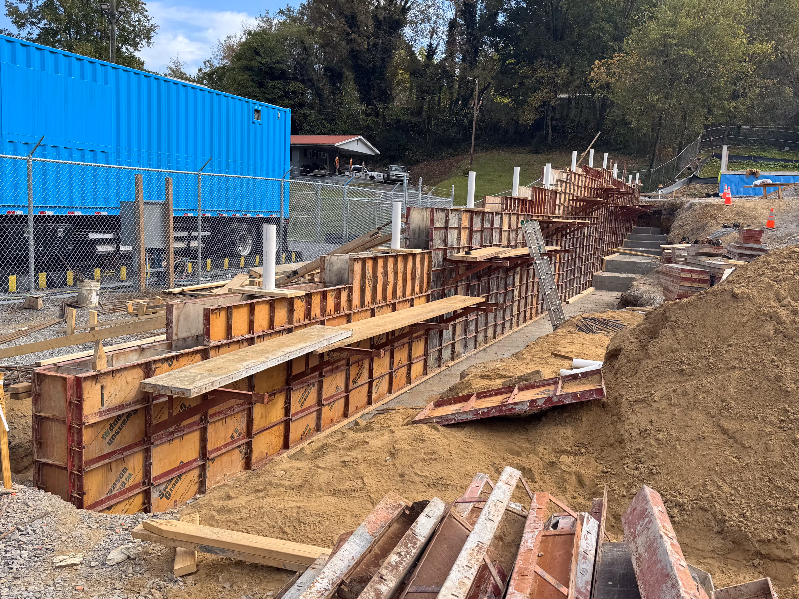 Construction site with wooden formwork for concrete wall and dirt piles beside a fenced area.