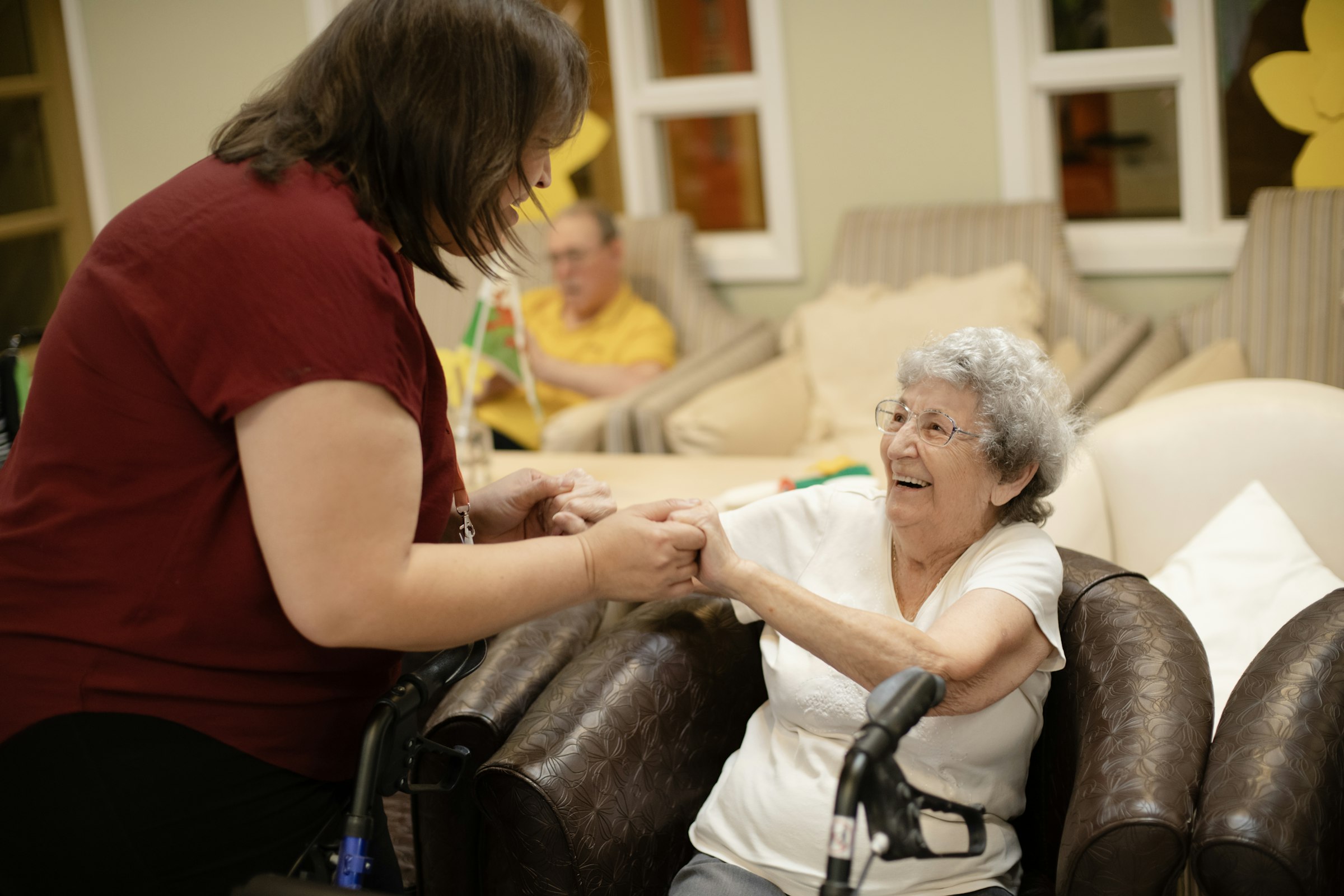 A home care worker holds hands with a smiling older woman in a bright living room