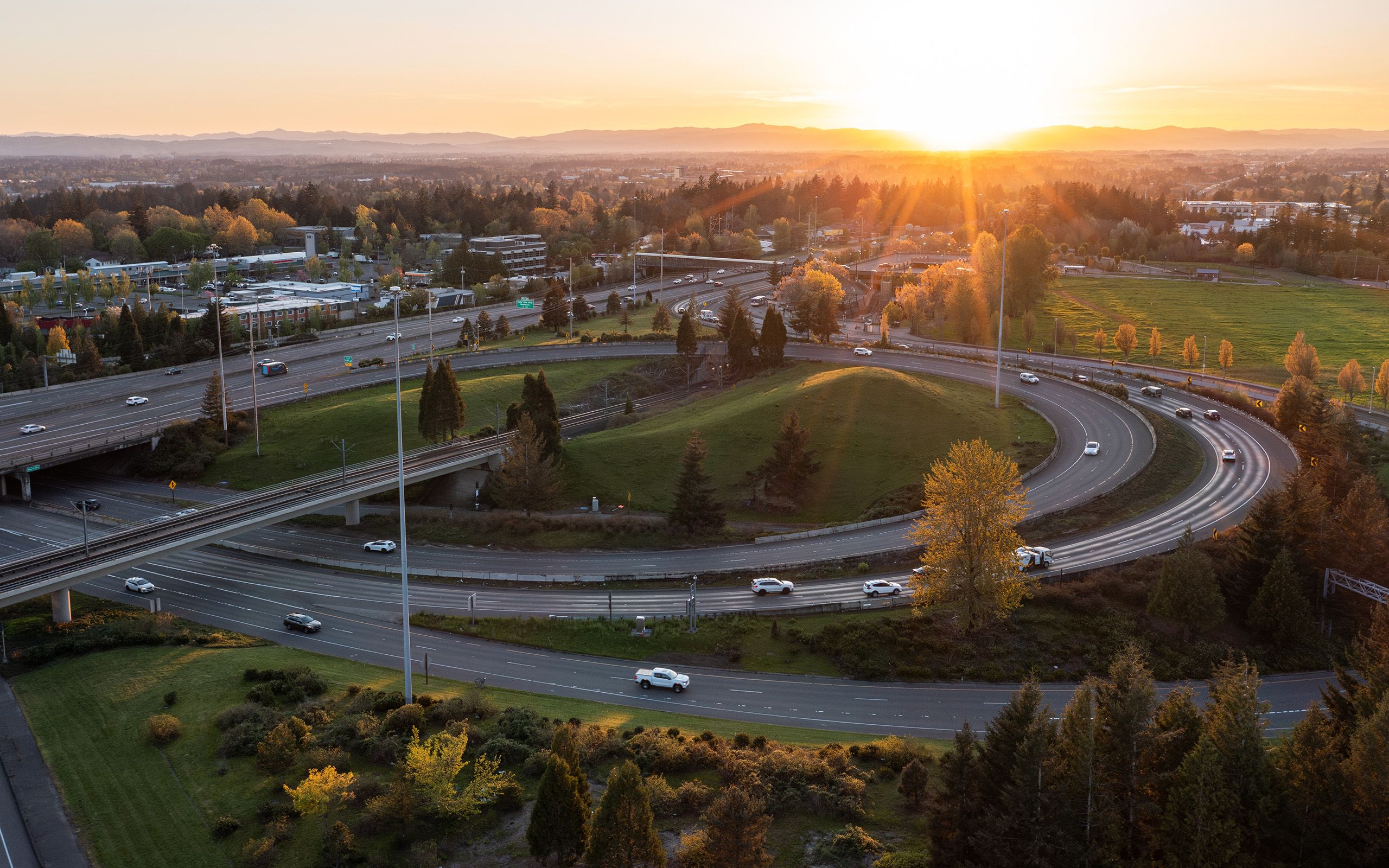 Aerial view of a freeway interchange at sunset with vehicles on curving ramps bordered by trees and green space