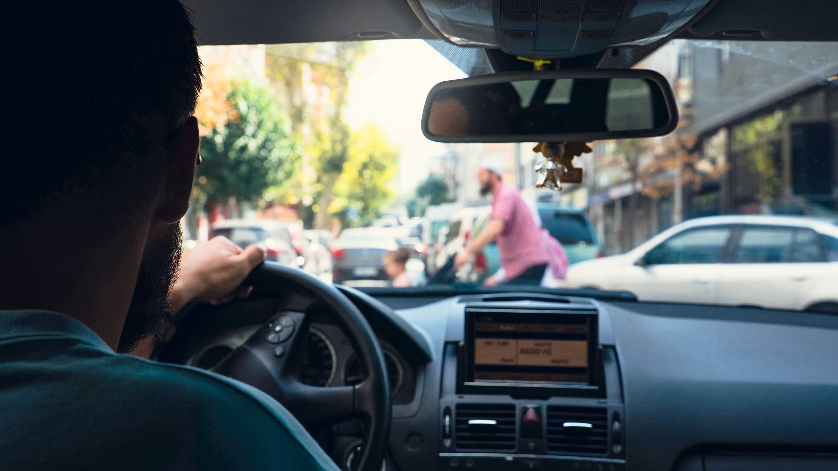 View from the driver seat through a car windshield on a busy city street, with pedestrians crossing and vehicles ahead