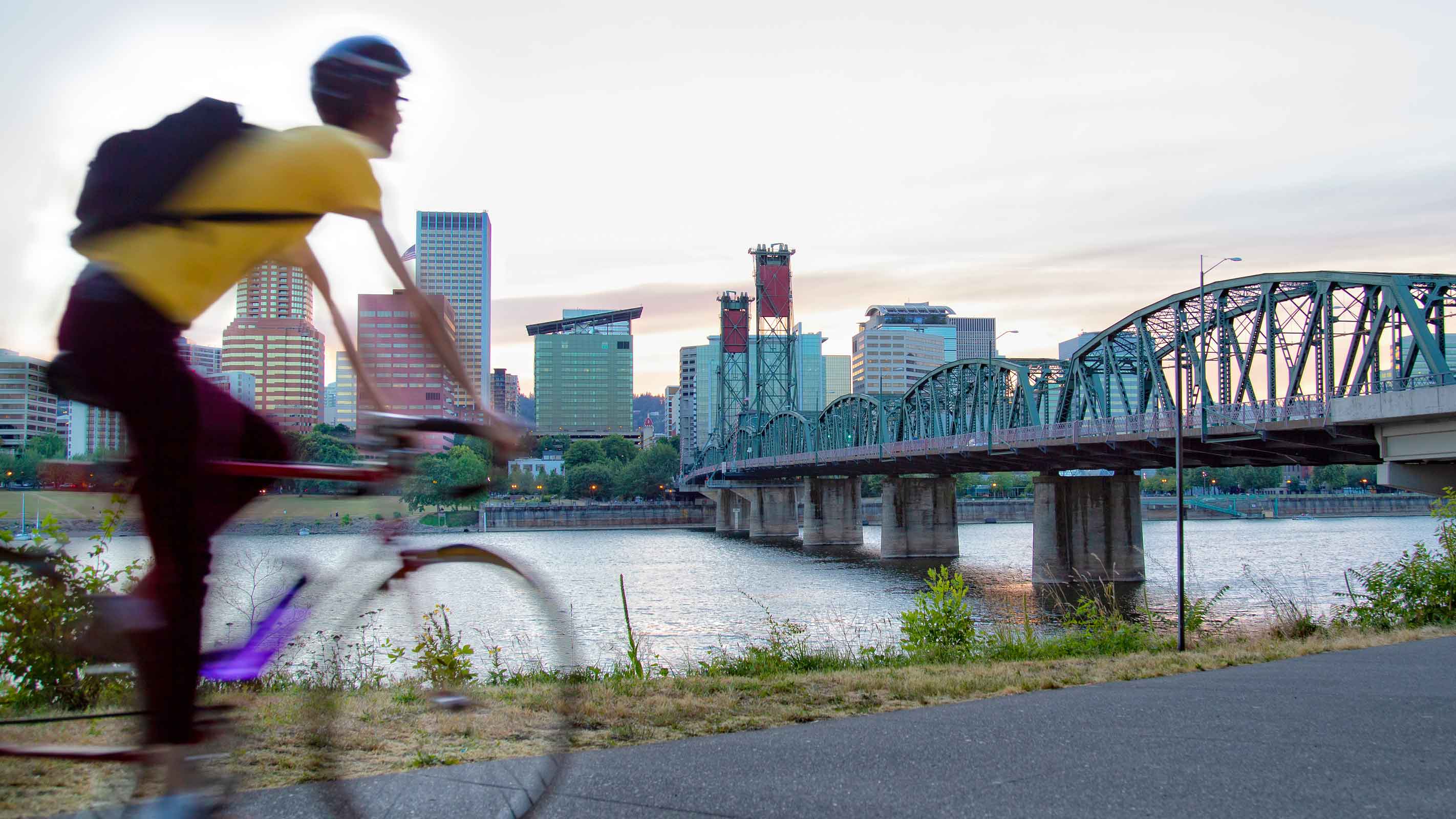 A person bikes along a riverfront path with the Hawthorne Bridge and the downtown Portland, Oregon skyline in the background