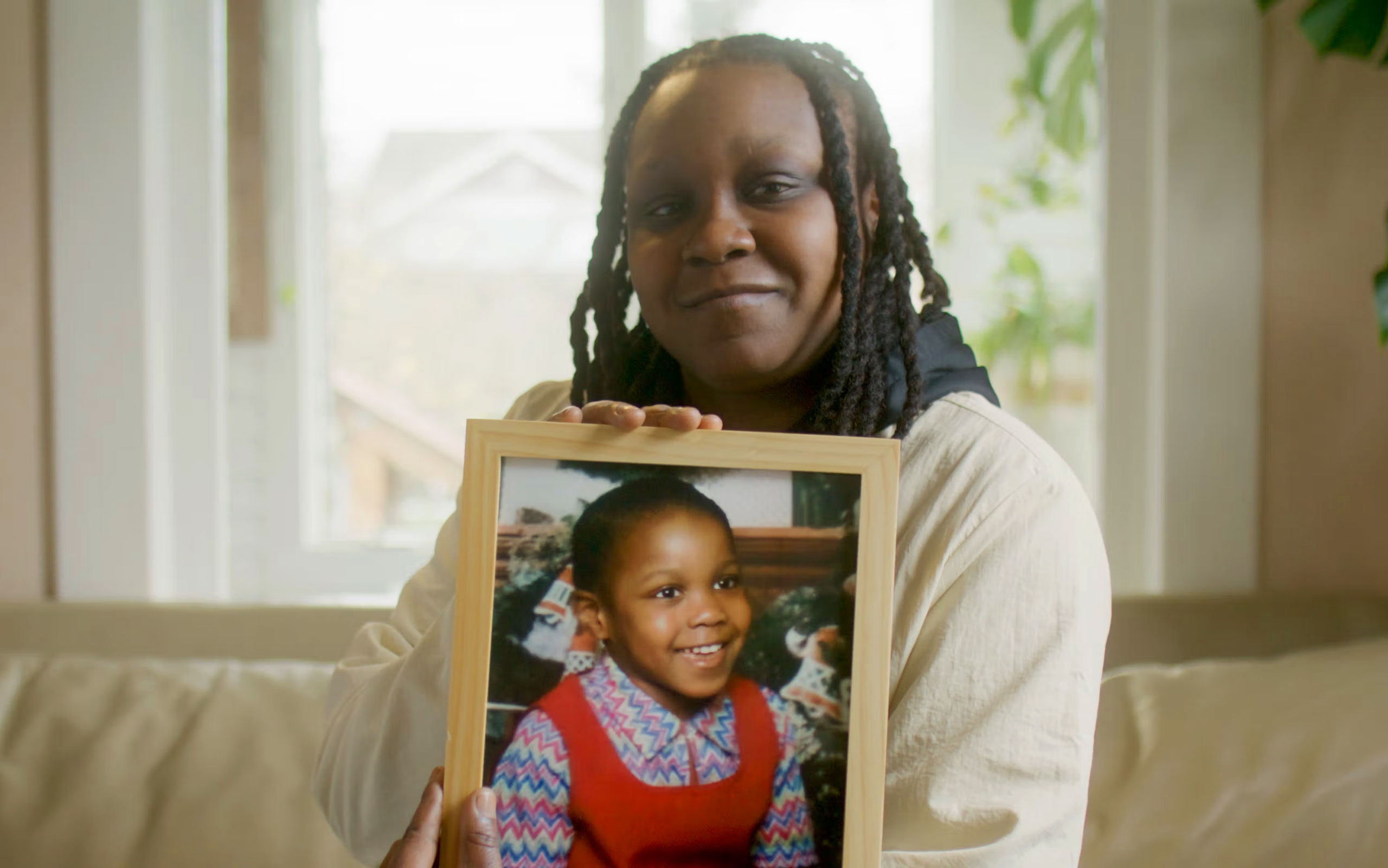 A smiling woman sits on a couch holding a framed photo of herself as a young child